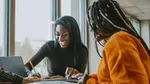 Two women working together at a desk, researching online and making notes