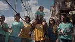 7 girls are between a fenced football pitch and buildings. Six are walking with one of them carrying a football. The smallest girl is being carried on the shoulders of the girl in the middle.