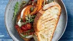 A delicious-looking open sandwich on a ceramic plate, featuring grilled bread topped with roasted pumpkin slices, cherry tomatoes, rocket, and pumpkin seeds. The vibrant colours and textures make it an appetising dish.
