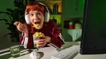 A person wearing large white headphones is eating instant noodles from a cup labelled 'Knorr' while sitting at a desk. The desk has a glass of water, a white computer keyboard, and a mouse and we can see the back of a computer monitor. The background includes shelves with books and plants, and the scene is lit with green ambient lighting.